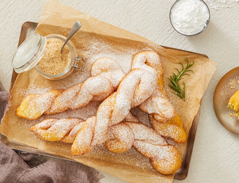 베이킹 트레이 위에 황금빛으로 튀겨진 꽈배기 도넛이 여러 개 놓여 있습니다. 도넛 위에는 하얀 슈가파우더가 고르게 뿌려져 있고, 옆에는 설탕가루와 빵가루가 담긴 작은 유리병이 놓여 있습니다. 도넛 옆에는 초록색 허브 가지가 장식되어 있으며, 전체적으로 따뜻하고 아늑한 분위기의 베이지색 천 위에 세팅되어 있습니다.