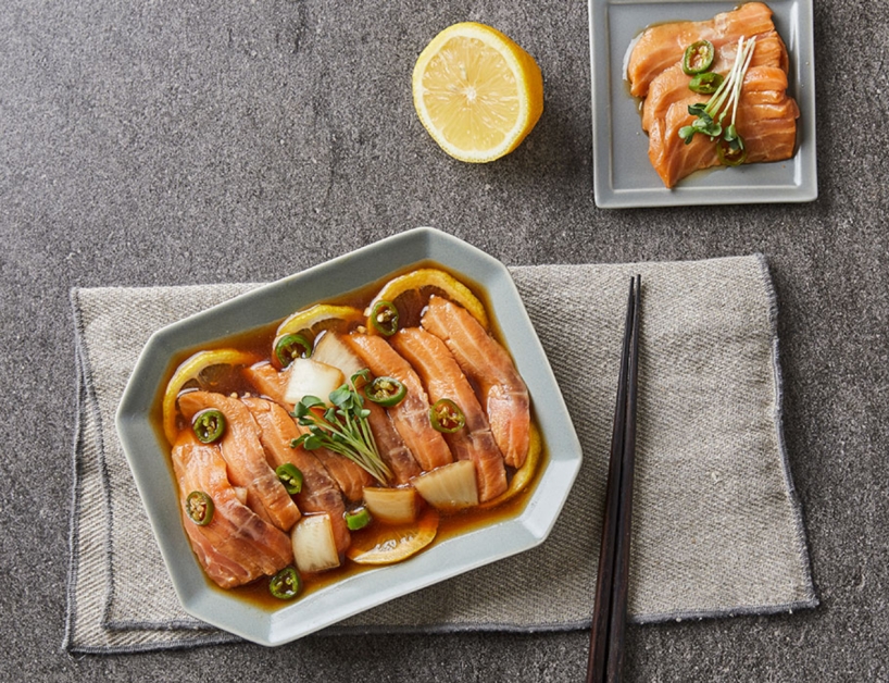 회색 천 위에 사각 접시에 담긴 연어회가 놓여 있습니다.
연어 위에는 얇게 썬 양파와 초록색 고추, 레몬 조각이 올려져 있으며, 옆에는 간장 소스가 담긴 작은 접시가 함께 배치되어 있습니다.
접시 오른쪽에는 젓가락이 가지런히 놓여 있습니다.